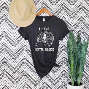 Op de afbeelding: Donkergrijs t-shirt met de witte tekst "I HAVE MENTAL ILLINOIS". De afbeelding toont een hersendiagram met de staat Illinois gemarkeerd. Een beige hoed hangt boven het shirt aan een houten hanger.