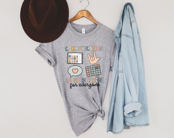 Puede incluir: Una camiseta gris jaspeado con el texto "COMMUNICATION looks DIFFERENT for everyone". La camiseta presenta ilustraciones coloridas de una tableta, una mano haciendo el signo "Te amo", una calculadora y un bocadillo con un corazón.