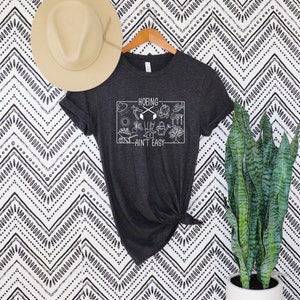 Op de afbeelding: Donkergrijs t-shirt met de tekst "HOEING AIN'T EASY" in een witte rechthoek, met tuingereedschap en elementen. Een beige hoed hangt boven het shirt. De achtergrond is een zwart-wit zigzagpatroon. Een plant in een pot staat rechts.