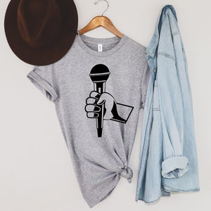 May include: A heather gray t-shirt featuring a black graphic of a hand holding a microphone. The shirt is hanging on a wooden hanger with a brown hat and a light blue denim jacket. The shirt has a crew neck and short sleeves.