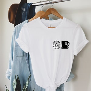Puede incluir: Camiseta blanca con un gráfico negro de una rosquilla y una taza de té. La rosquilla tiene un agujero y chispas. La taza de té con una bolsita descansa sobre un platillo. La camiseta está colgada en una percha de madera.