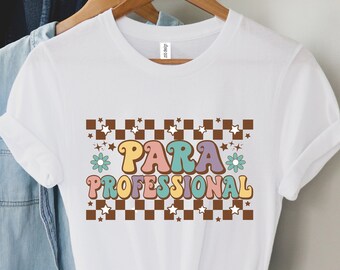 May include: White t-shirt featuring the text "PARA PROFESSIONAL" in a retro design with a brown and white chequered pattern, stars, and flowers. The shirt is displayed on a wooden hanger, showcasing a vintage aesthetic.