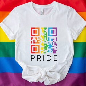 May include: White t-shirt with a colorful QR code design in rainbow colors. The word "PRIDE" is printed below the QR code in black. The shirt is displayed on a rainbow flag background.