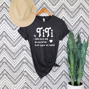 May include: Dark gray t-shirt with the word "TiTi" in white script. The shirt includes a definition: "Another term for aunty like a mom but Cooler." A beige hat hangs above the shirt, and a tall plant is to the right.
