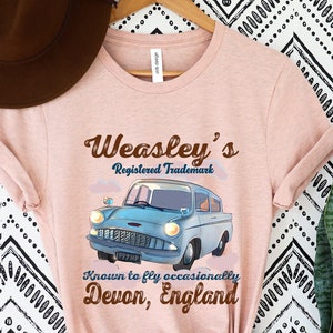 May include: A light pink t-shirt featuring a blue vintage car graphic with the text "Weasley's Registered Trademark." Additional text reads "Known to fly occasionally, Devon, England." The shirt is hanging on a wooden hanger.