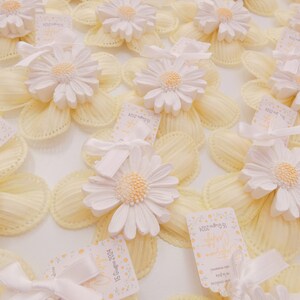 Può includere: Una collezione di scatole portaconfetti gialle e bianche con un fiore di margherita bianco in cima. Ogni scatola ha un piccolo cartellino con il testo "2023 Wedding" e una data.