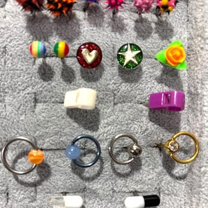 Pu&ograve; includere: Una collezione di gioielli per il corpo, tra cui orecchini e piercing al naso, esposta su una superficie grigia. I gioielli presentano vari design, come punte, cuori, stelle e forme geometriche. I colori includono rosso, arancione, blu, rosa, verde e giallo.