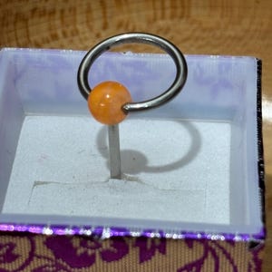 Pu&ograve; includere: Un anello captive bead di colore argento con una perla arancione. L'anello &egrave; esposto all'interno di una piccola scatola aperta con un esterno viola e a motivi floreali. La scatola ha un interno bianco.