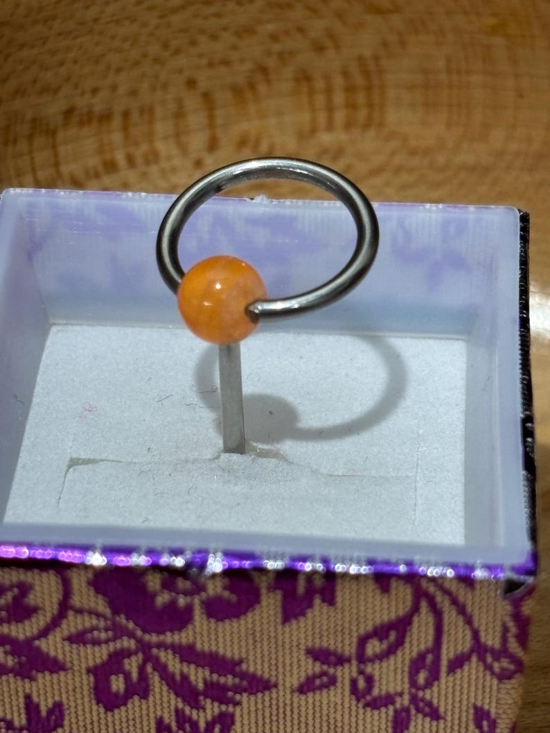 Pu&ograve; includere: Un anello captive bead di colore argento con una perla arancione. L'anello &egrave; esposto all'interno di una piccola scatola aperta con un esterno viola e a motivi floreali. La scatola ha un interno bianco.