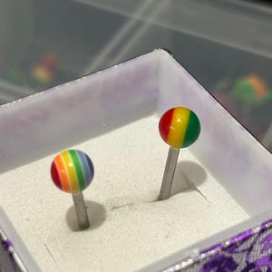 Pu&ograve; includere: Due orecchini a lobo arcobaleno con perni d'argento sono esposti in una scatola decorativa. Gli orecchini presentano un design sferico con bande rosse, arancioni, gialle, verdi, blu e viola. La scatola ha un interno bianco e un esterno viola a motivi floreali.