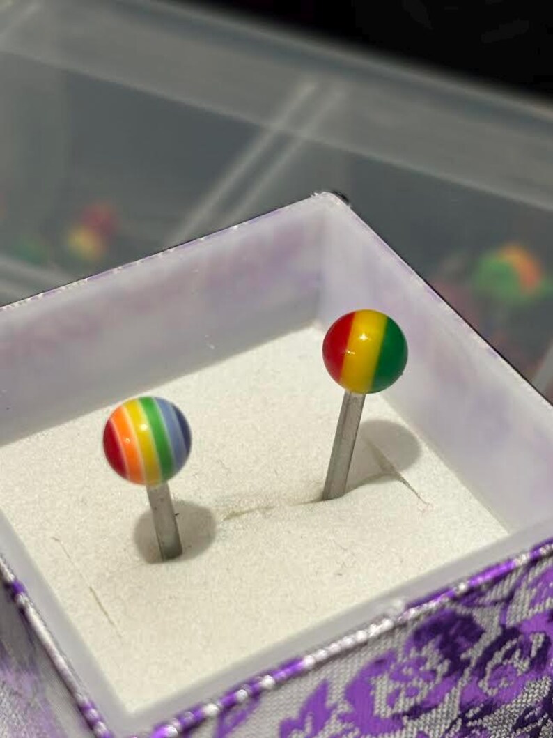 Pu&ograve; includere: Due orecchini a lobo arcobaleno con perni d'argento sono esposti in una scatola decorativa. Gli orecchini presentano un design sferico con bande rosse, arancioni, gialle, verdi, blu e viola. La scatola ha un interno bianco e un esterno viola a motivi floreali.