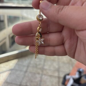 K&ouml;nnte beinhalten: Ein goldfarbener Bauchnabelpiercing mit einem klaren, runden Kristall an der Oberseite, einem Anh&auml;nger in Blumenform mit klaren Steinen im Marquise-Schliff und zwei feinen Goldketten, die f&uuml;r zus&auml;tzliche Bewegung an der Blume h&auml;ngen.