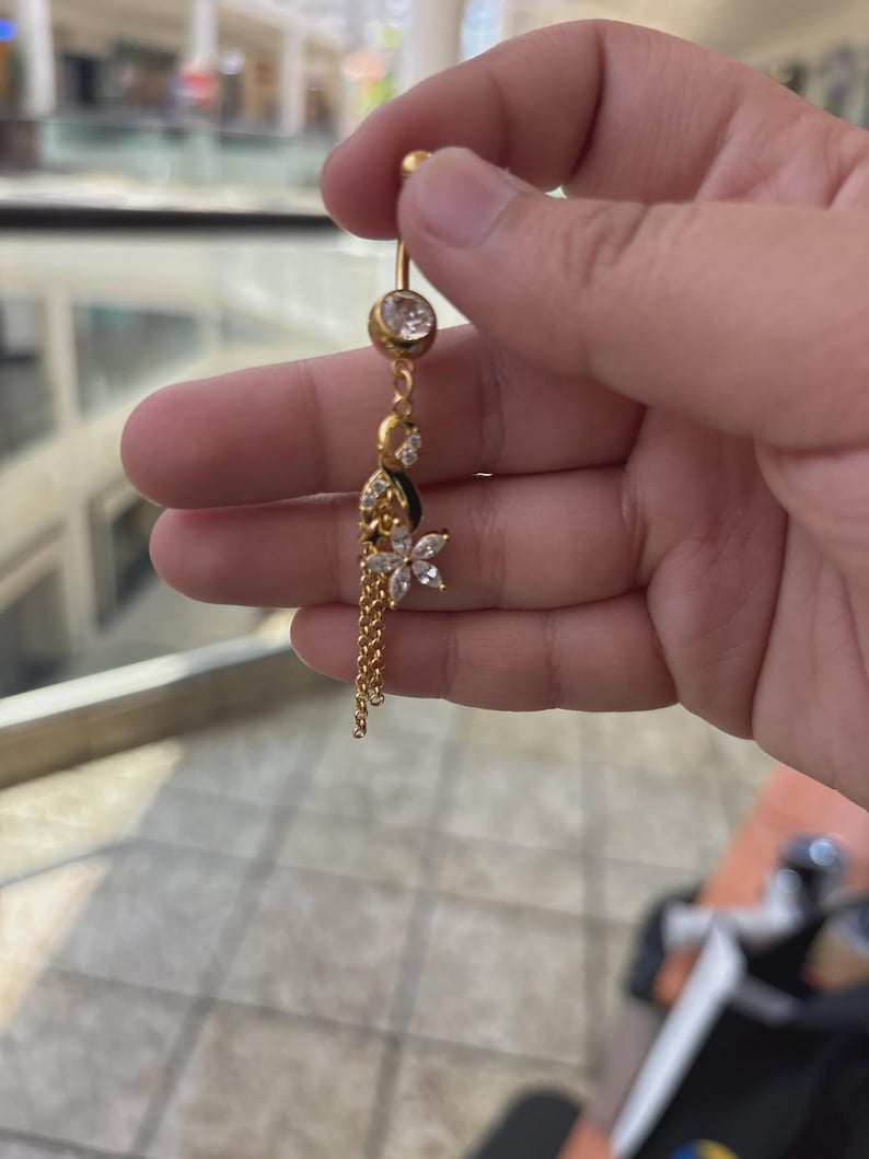 K&ouml;nnte beinhalten: Ein goldfarbener Bauchnabelpiercing mit einem klaren, runden Kristall an der Oberseite, einem Anh&auml;nger in Blumenform mit klaren Steinen im Marquise-Schliff und zwei feinen Goldketten, die f&uuml;r zus&auml;tzliche Bewegung an der Blume h&auml;ngen.