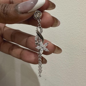 K&ouml;nnte beinhalten: Ein silberfarbener Bauchnabelpiercing mit einer oberen Kugel, einem gr&ouml;&szlig;eren klaren Kristallakzent und einem h&auml;ngenden Anh&auml;nger. Der Anh&auml;nger besteht aus einem Unendlichkeitssymbol mit kleinen klaren Steinen, einer sechsbl&auml;ttrigen Blume und einer Kette.