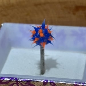 Pu&ograve; includere: Una puntina decorativa con una testa sferica e appuntita. La testa &egrave; una combinazione di blu e arancione, montata su un perno di metallo color argento. La puntina &egrave; esposta all'interno di una piccola scatola con un bordo a motivi viola e oro.