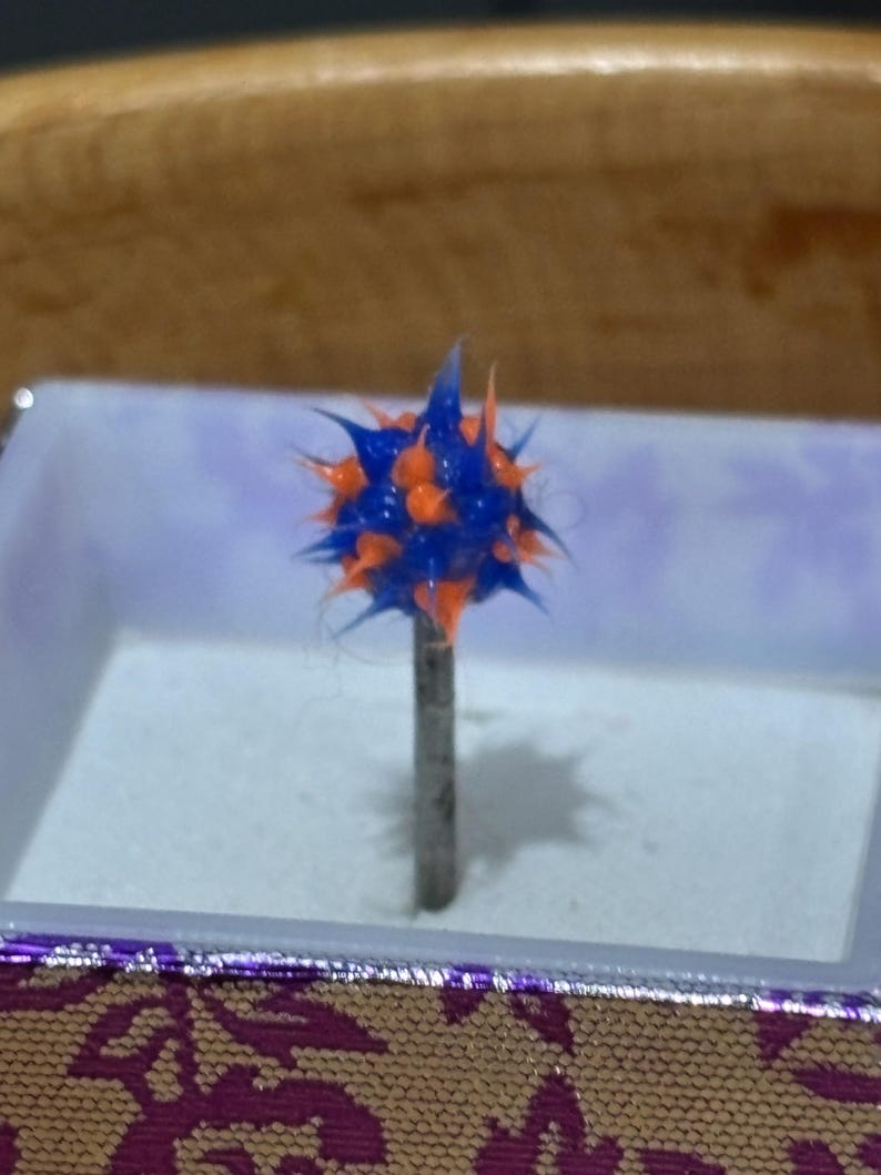 Pu&ograve; includere: Una puntina decorativa con una testa sferica e appuntita. La testa &egrave; una combinazione di blu e arancione, montata su un perno di metallo color argento. La puntina &egrave; esposta all'interno di una piccola scatola con un bordo a motivi viola e oro.