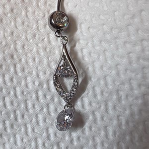 K&ouml;nnte beinhalten: Ein silberfarbener Bauchnabelpiercing mit einem gebogenen Stab und einem runden, klaren Stein an der Basis. Ein tropfenf&ouml;rmiger Anh&auml;nger h&auml;ngt an dem Stab, besetzt mit kleinen klaren Steinen und einem runden Mittelstein, der in einem ovalen Stein endet.