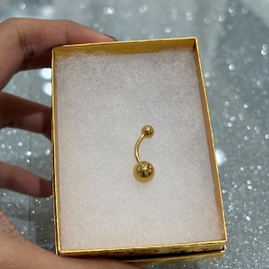May include: A gold-colored belly button ring with a curved barbell design. The jewelry is displayed inside a small, gold-colored box with a white, cotton-like interior.