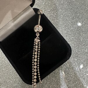 May include: A silver belly button ring with a dangling chain of small, clear rhinestones. The ring is displayed in an open black jewelry box against a sparkly silver background. The ring has a small ball at the top and a larger, round rhinestone at the center.