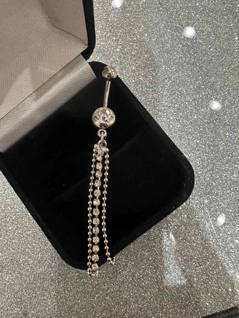 May include: A silver belly button ring with a dangling chain of small, clear rhinestones. The ring is displayed in an open black jewelry box against a sparkly silver background. The ring has a small ball at the top and a larger, round rhinestone at the center.