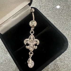 May include: Silver-toned belly button ring with a fleur-de-lis design. The ring features a curved barbell with a small ball at the top and a larger, round gem in the middle. A fleur-de-lis pendant with clear gems hangs below, with a teardrop gem at the bottom.