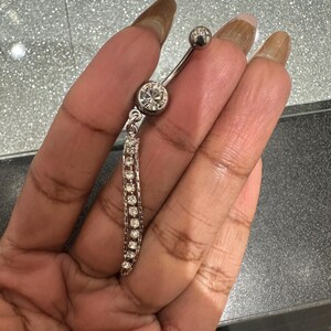 May include: A silver belly button ring with a dangling chain of small, clear gemstones. The curved barbell has a large, round gemstone at the bottom and a smaller, round gemstone at the top.