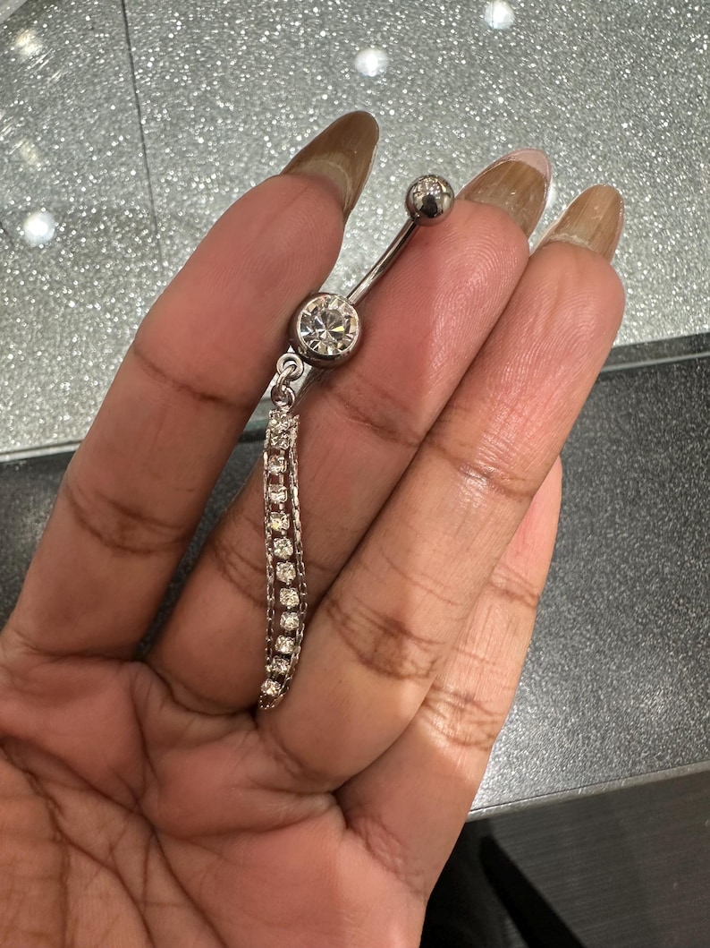 May include: A silver belly button ring with a dangling chain of small, clear gemstones. The curved barbell has a large, round gemstone at the bottom and a smaller, round gemstone at the top.