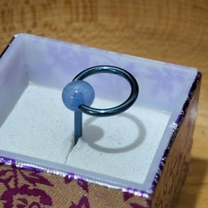 Pu&ograve; includere: Un anello con perla blu e cerchio e asta in metallo blu scuro, presentato in un piccolo cofanetto decorativo con un motivo floreale viola e oro. L'anello &egrave; posto su uno sfondo bianco.
