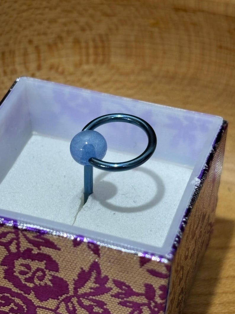 Pu&ograve; includere: Un anello con perla blu e cerchio e asta in metallo blu scuro, presentato in un piccolo cofanetto decorativo con un motivo floreale viola e oro. L'anello &egrave; posto su uno sfondo bianco.