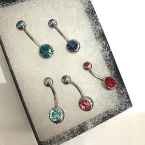 May include: A collection of six belly button rings with curved silver-toned bars and colorful, round gemstones. The gemstones are in shades of teal, blue, pink, and red. The jewelry is displayed in a black box with white padding.