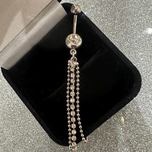 May include: A silver belly button ring with a large, clear, round gem at the center and a smaller gem on top. Three strands of small, sparkling gems dangle from the center gem. The jewelry is displayed in a black velvet box.