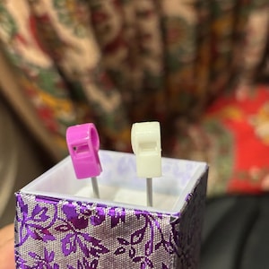 Pu&ograve; includere: Una piccola scatola decorativa con un motivo floreale viola e argento. All'interno, sono esposti due fischietti in miniatura: uno rosa e uno bianco. La scatola &egrave; tenuta in mano da una persona, con uno sfondo sfocato.