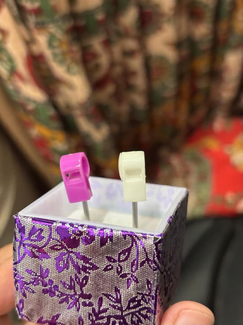 Pu&ograve; includere: Una piccola scatola decorativa con un motivo floreale viola e argento. All'interno, sono esposti due fischietti in miniatura: uno rosa e uno bianco. La scatola &egrave; tenuta in mano da una persona, con uno sfondo sfocato.