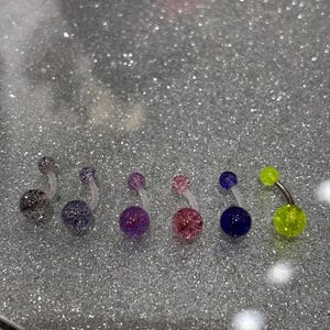 May include: A collection of belly button rings with various colors and glittery finishes. The rings include a silver, purple, pink, blue, and neon green. Each ring has a curved barbell with a clear, flexible shaft.
