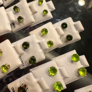 May include: An assortment of stud earrings with round, green gemstones. The earrings are displayed on small white cards, some of which have the text "CUBIC ZIRCONIA" printed on them. The gemstones vary in shade from light to dark green.