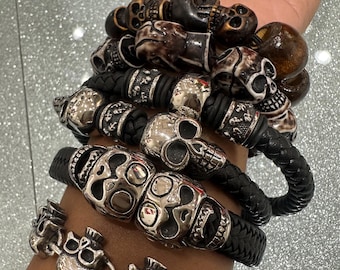 May include: Several bracelets with skull motifs. The bracelets are crafted from materials such as silver-toned metal, dark brown wood, and black leather. The skull designs vary, offering a striking and edgy look for the wrist.