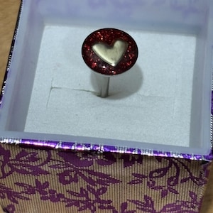 Pu&ograve; includere: Un anello con una parte superiore ovale rossa glitterata con un design a cuore dorato. L'anello &egrave; esposto all'interno di una scatola decorativa con un motivo floreale viola e oro.