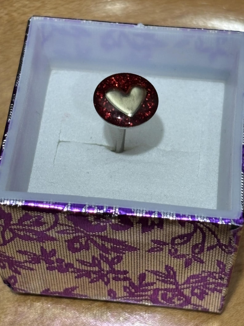 Pu&ograve; includere: Un anello con una parte superiore ovale rossa glitterata con un design a cuore dorato. L'anello &egrave; esposto all'interno di una scatola decorativa con un motivo floreale viola e oro.