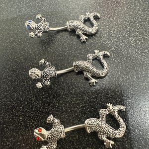 May include: Three silver belly button rings with a lizard design. The lizards have jeweled eyes, one blue, one red, and one clear.