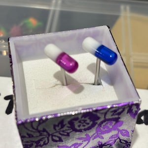 Pu&ograve; includere: Un paio di gemelli a forma di capsule, uno viola e bianco, l'altro blu e bianco, esposti in una scatola decorativa con un motivo floreale viola.