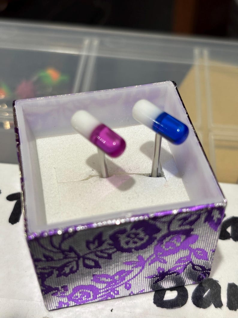 Pu&ograve; includere: Un paio di gemelli a forma di capsule, uno viola e bianco, l'altro blu e bianco, esposti in una scatola decorativa con un motivo floreale viola.