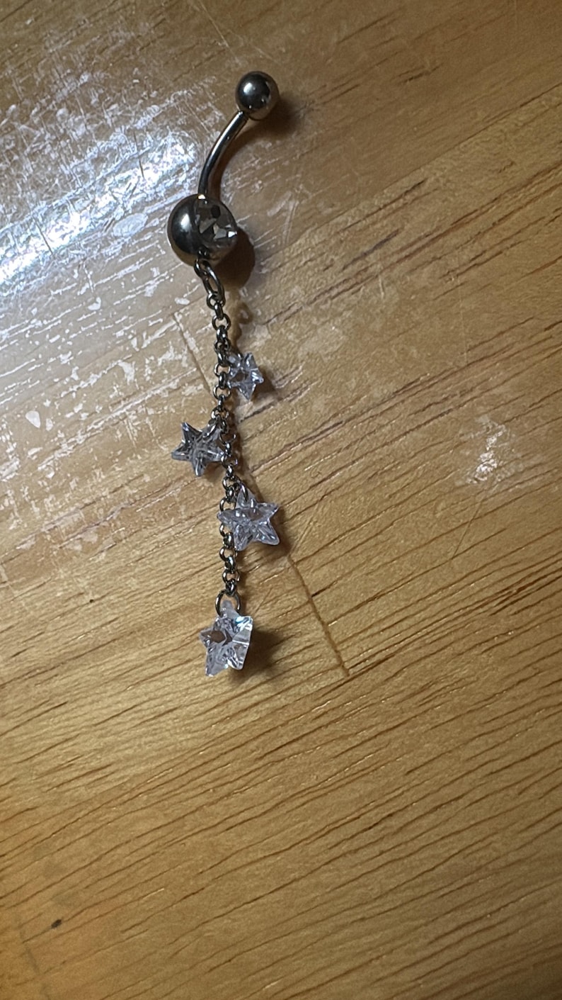 K&ouml;nnte beinhalten: Ein silberfarbener Bauchnabelpiercing mit einem gebogenen Stab und einem runden, klaren Stein an der Basis. Eine feine silberne Kette h&auml;ngt daran herab, verziert mit vier kleinen, klaren, sternf&ouml;rmigen Kristallen, die in einer Reihe h&auml;ngen.