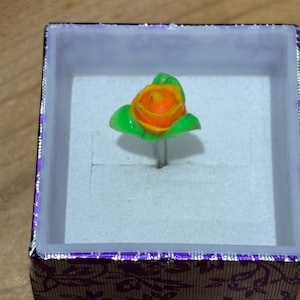 Pu&ograve; includere: Anello fatto a mano con una mini rosa arancione con foglie verdi. Il design floreale &egrave; montato su una fascia color argento e presentato in una scatola viola decorativa con un motivo floreale.