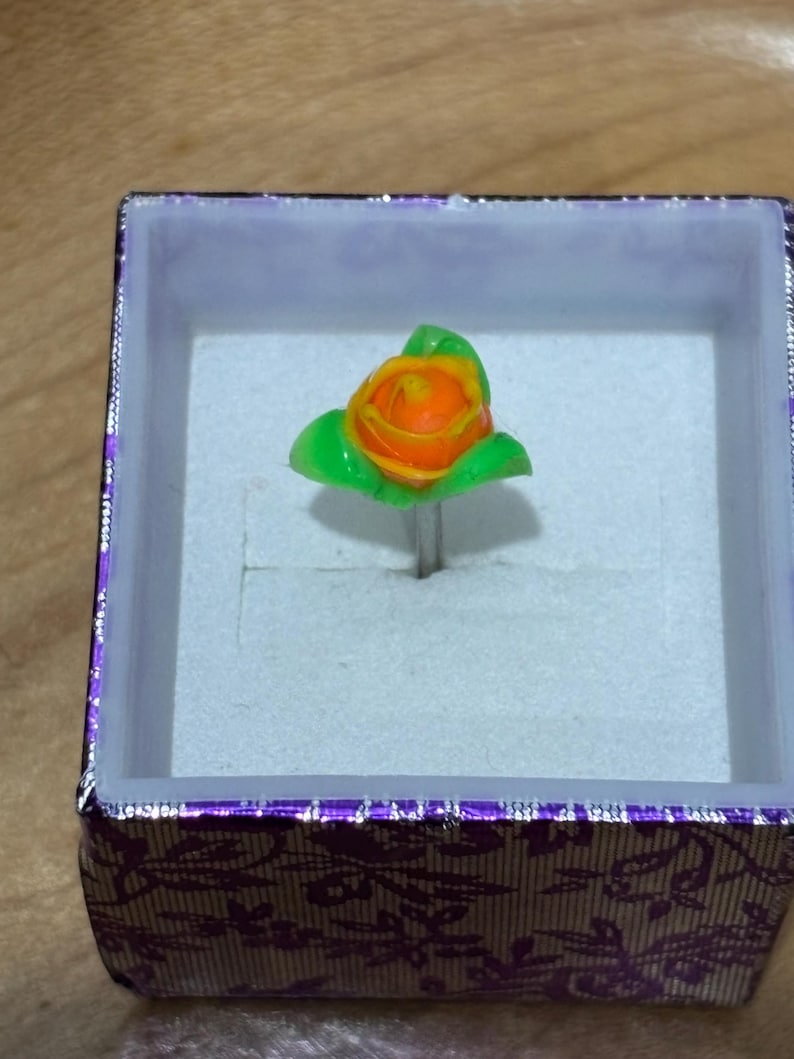 Pu&ograve; includere: Anello fatto a mano con una mini rosa arancione con foglie verdi. Il design floreale &egrave; montato su una fascia color argento e presentato in una scatola viola decorativa con un motivo floreale.