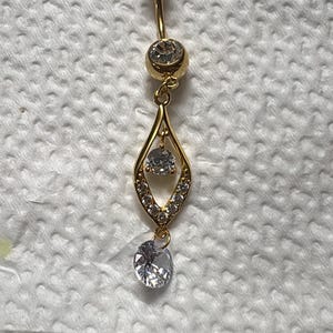 K&ouml;nnte beinhalten: Ein goldfarbener Bauchnabelpiercing mit einem gebogenen Stab, einer oberen Kugel und einem gr&ouml;&szlig;eren unteren Stein. Daran befestigt ist ein tropfenf&ouml;rmiger Anh&auml;nger, der mit kleinen klaren Kristallen und einem runden Stein an der Basis verziert ist.