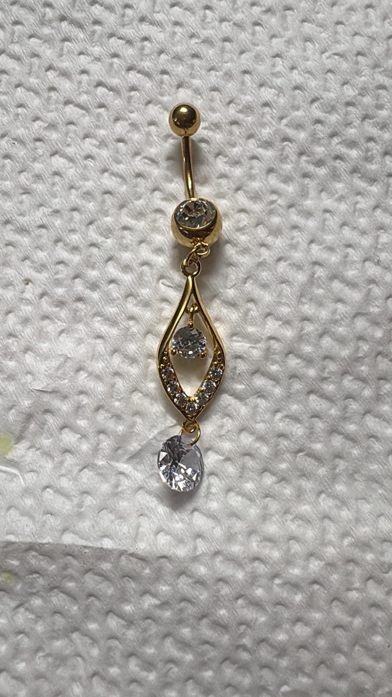 K&ouml;nnte beinhalten: Ein goldfarbener Bauchnabelpiercing mit einem gebogenen Stab, einer oberen Kugel und einem gr&ouml;&szlig;eren unteren Stein. Daran befestigt ist ein tropfenf&ouml;rmiger Anh&auml;nger, der mit kleinen klaren Kristallen und einem runden Stein an der Basis verziert ist.