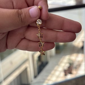 K&ouml;nnte beinhalten: Ein goldfarbener Bauchnabelpiercing mit einer oberen Kugel, einem gr&ouml;&szlig;eren runden klaren Kristall und einer h&auml;ngenden Kette mit drei kleinen sternf&ouml;rmigen Anh&auml;ngern. Jeder Sternanh&auml;nger ist teilweise mit winzigen klaren Kristallen besetzt.