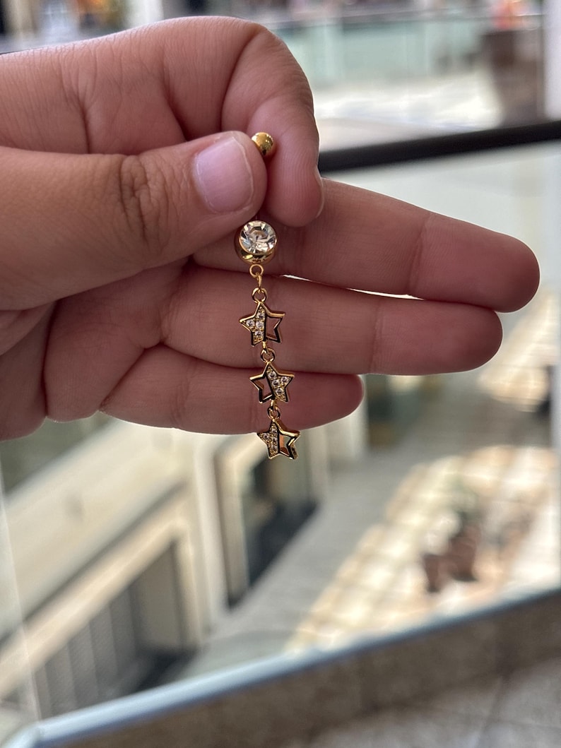 K&ouml;nnte beinhalten: Ein goldfarbener Bauchnabelpiercing mit einer oberen Kugel, einem gr&ouml;&szlig;eren runden klaren Kristall und einer h&auml;ngenden Kette mit drei kleinen sternf&ouml;rmigen Anh&auml;ngern. Jeder Sternanh&auml;nger ist teilweise mit winzigen klaren Kristallen besetzt.