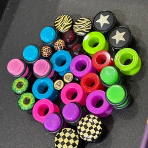 May include: A collection of circular ear plugs in various colors and patterns, including neon green, bright blue, hot pink, and purple. Some feature designs like black and white checkerboards, animal prints, stars, skulls, and realistic green eyes.