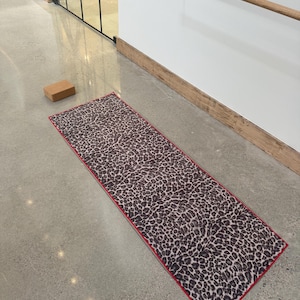 May include: A rectangular yoga mat with a leopard print pattern in shades of brown and tan, outlined with a red border. A wooden yoga block is visible nearby. The mat is on a concrete floor.